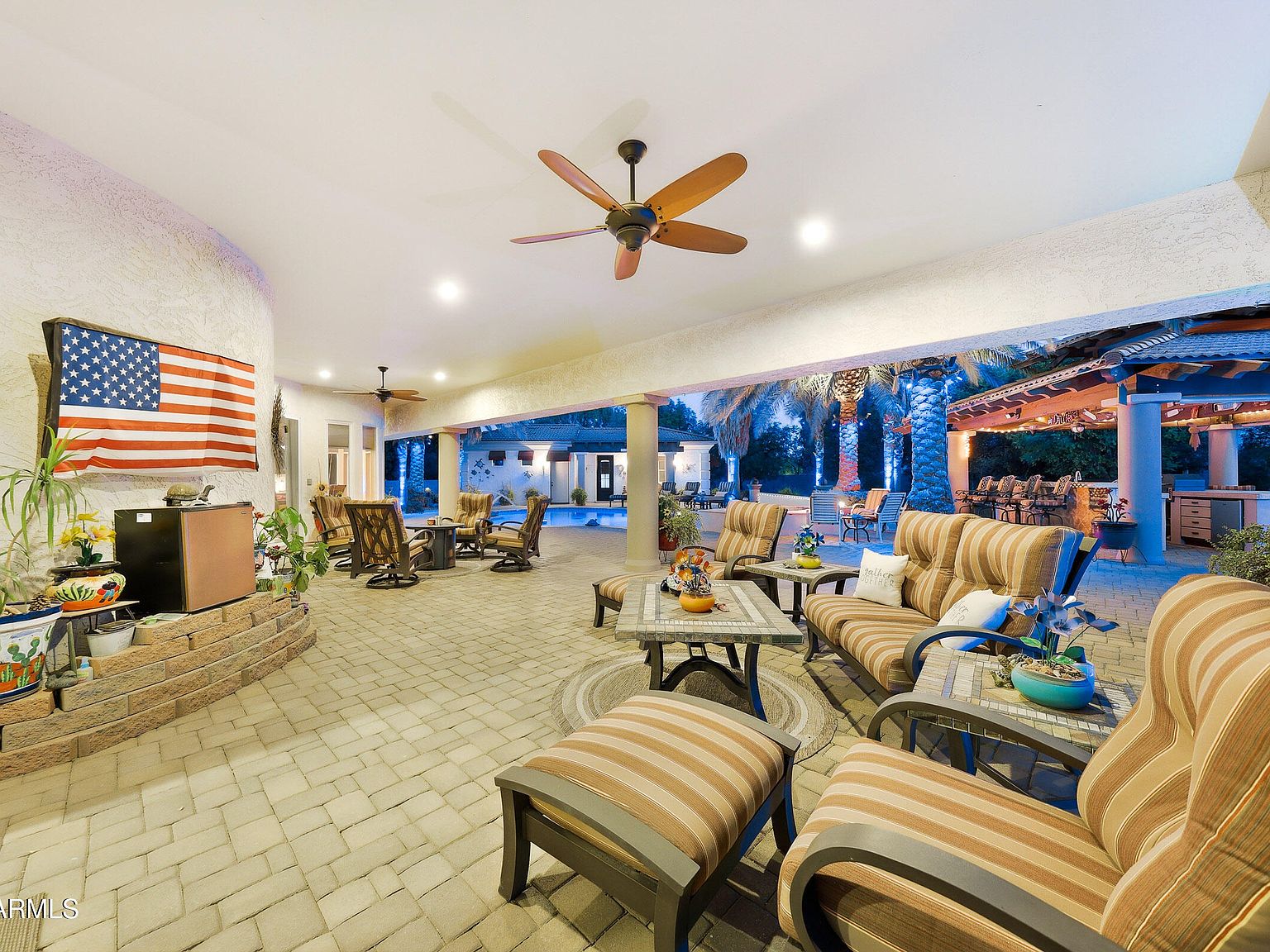 The covered patio at twilight with a ceiling fan, striped lounge seating, pavers underfoot, and a long view across the lit pool toward the gazebo