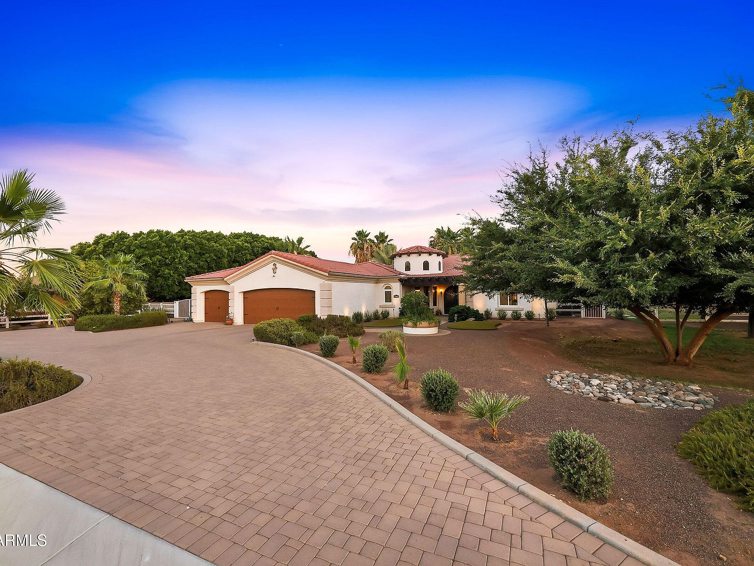 The front facade at twilight viewed across the paver motor court, with mature fan palms flanking the main house and the detached casita off to the left