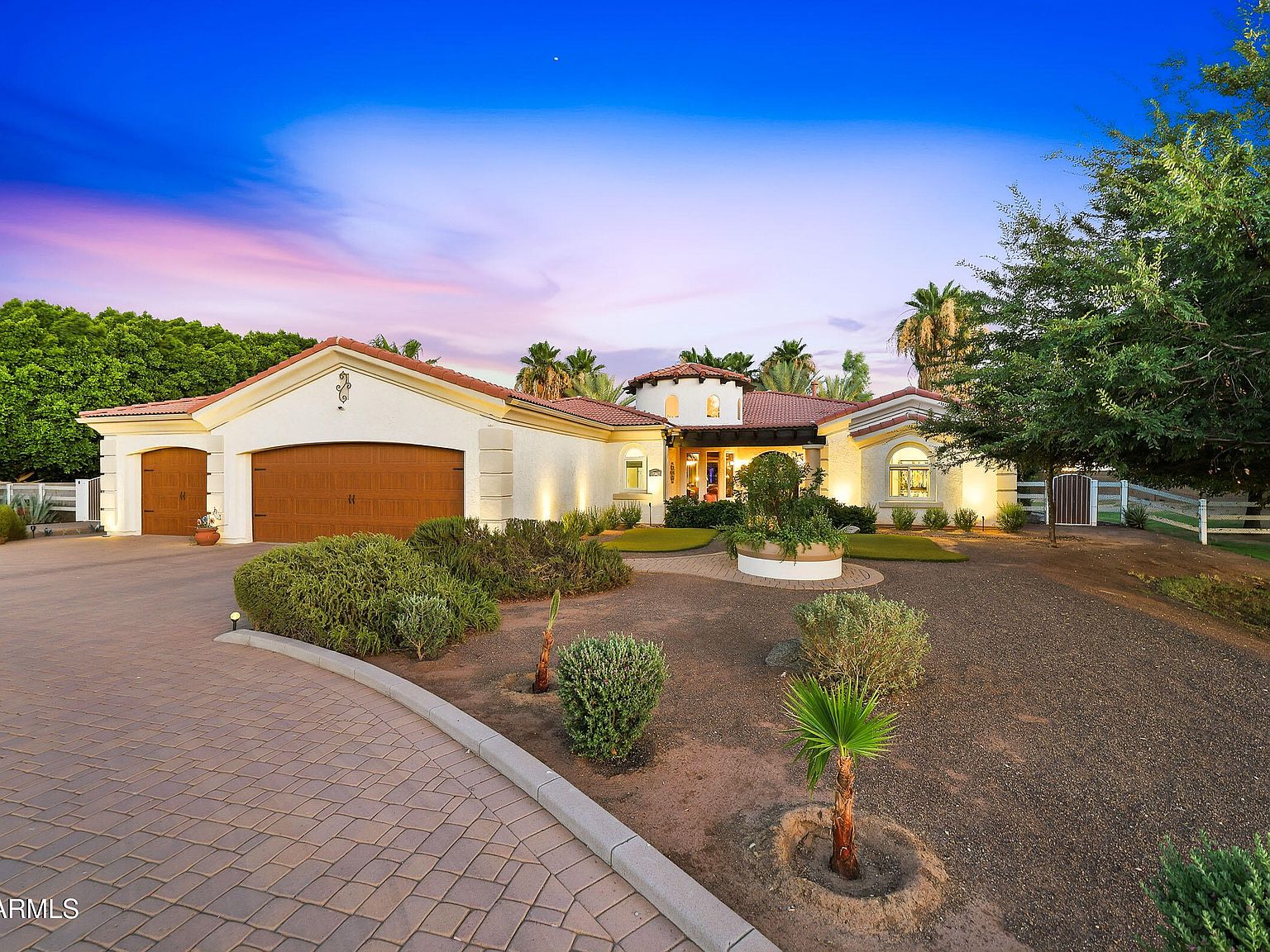 Front elevation of 17347 W Lamar Road at twilight, a Spanish-style estate with tile-roof eaves, a lit entry, and a paver motor court framed by date palms under a pink and blue Sonoran sky.
